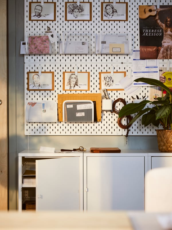 White SKÅDIS pegboards holding sketches, envelopes, a white SKÅDIS magazine holder and other items are attached to a wall.