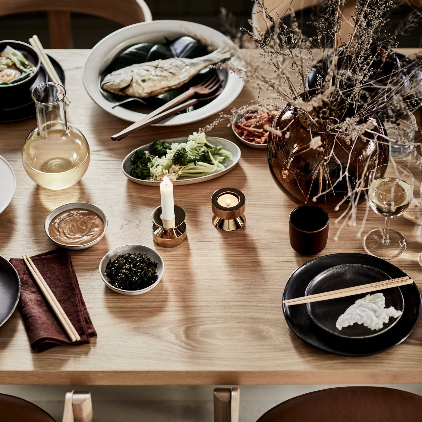 Herbstlich gedeckter Tisch mit der GLADELIG Serie in Dunkelgrau, STORSINT Gläsern, STOCKHOLM 2025 Stoffservietten in Dunkelgrün und Dunkelrot, STOCKHOLM 2025 Kerzenhaltern und Vase mit Trockenblumen.