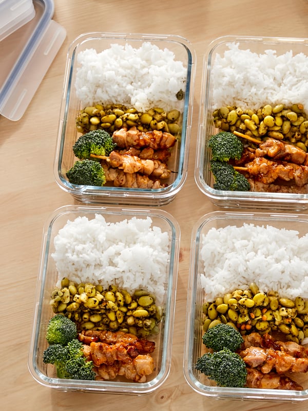 Four glass food containers filled with chicken skewers, rice, beans and broccoli stand on a wooden kitchen counter.