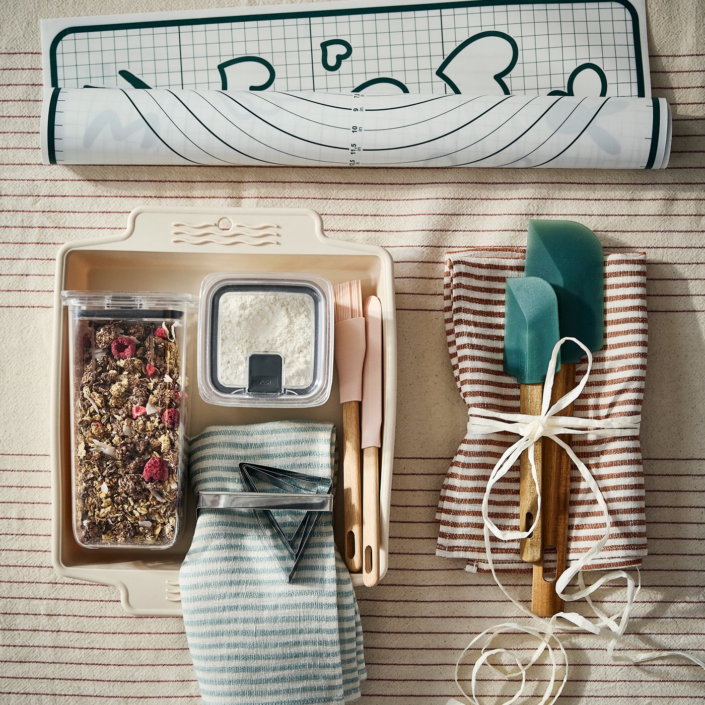One gift bundle of a tea towel and baking spatulas beside a BAKTIDER baking sheet filled with two food storage containers.