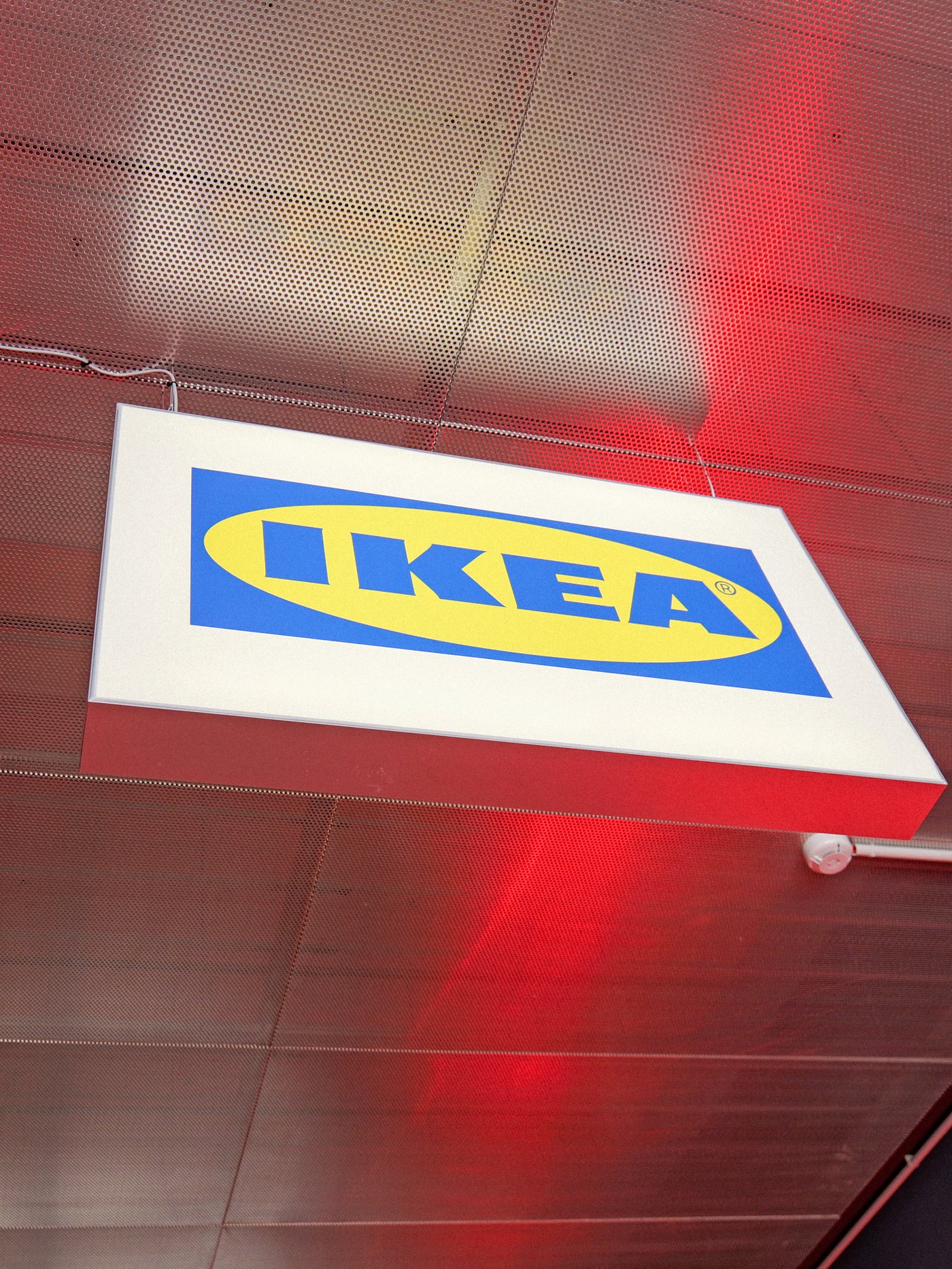 Insegna luminosa IKEA con logo blu e giallo sospesa al soffitto di un ambiente interno illuminato di rosso.