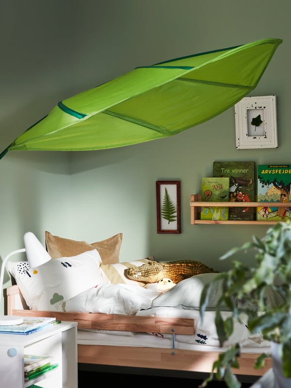 A corner of a child’s room with a SNIGLAR bed, a wall shelf with books and a large, leaf-shaped, green LÖVA bed canopy above.