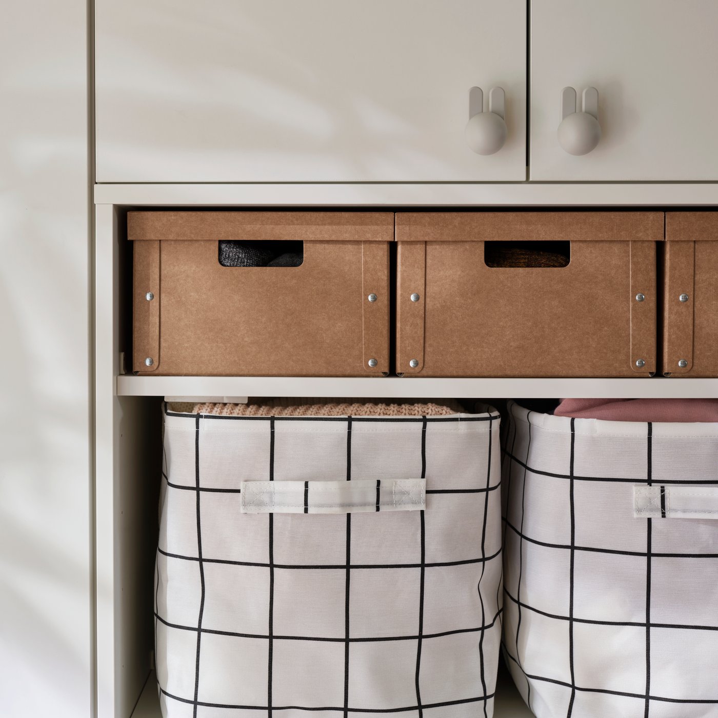 Étagère où se trouvent de grandes boîtes avec couvercles GÄRDESGÅRD au-dessus de grandes boîtes en tissu à motifs, dans une entrée.