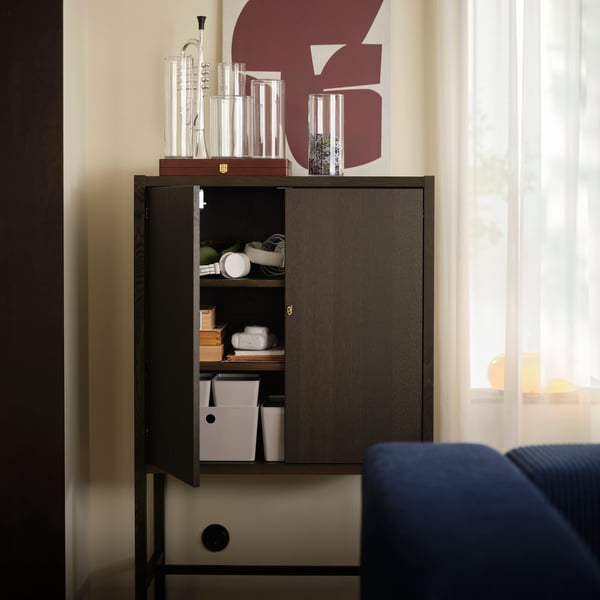 En vardagsrumsvägg med ett mörkbrunt STOCKHOLM 2025 skåp som rymmer olika accessoarer, bredvid ett fönster med skira gardiner.
