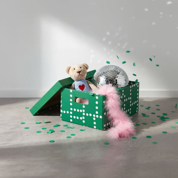 En grön förvaringslåda fylld med roliga festartiklar, inklusive en nallebjörn, en glänsande discokula och en rosa fjäderboa, med grön konfetti utspridd på golvet.