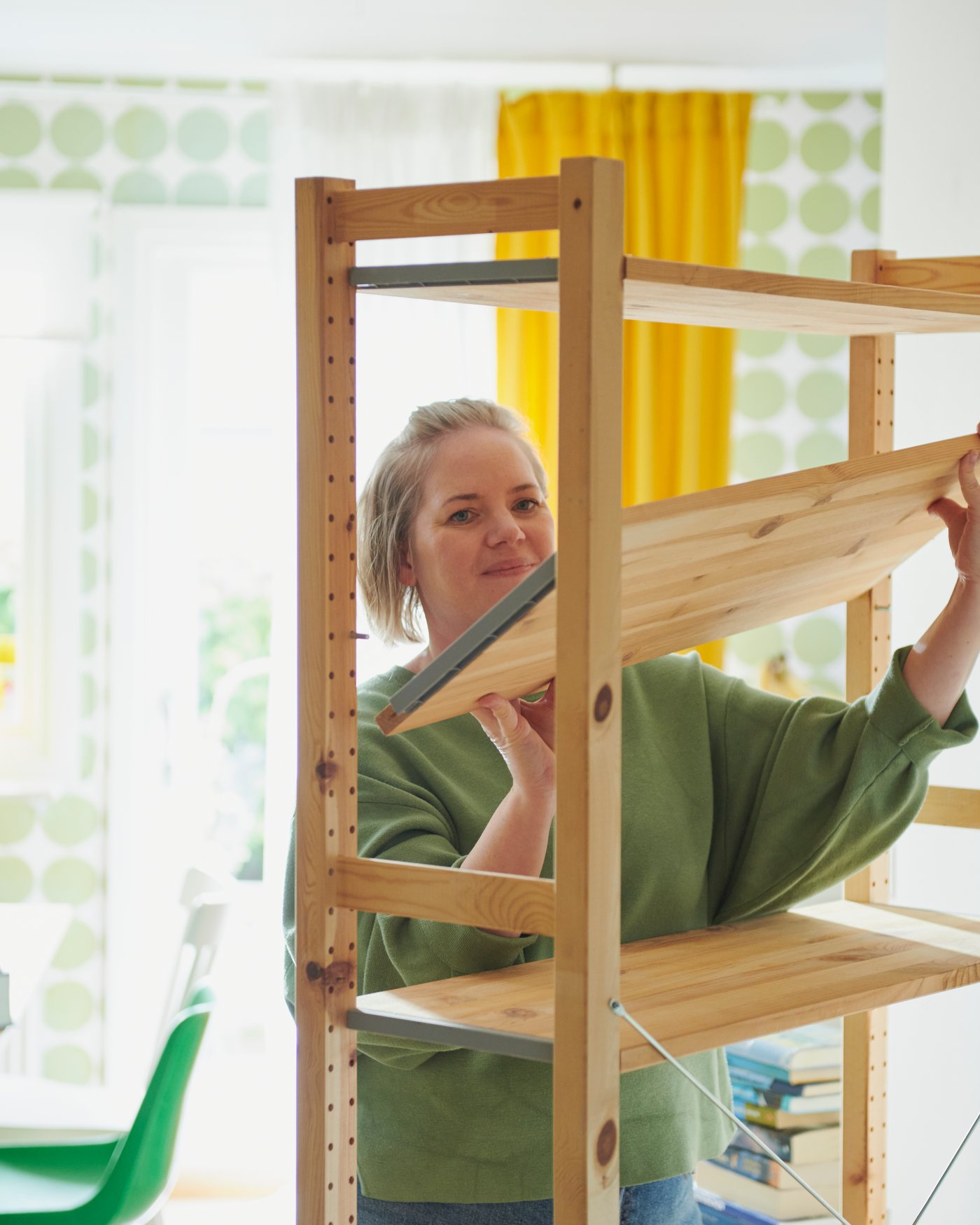 A person is assembling a pine IVAR shelving unit in a light-filled room with patterned wallpaper.