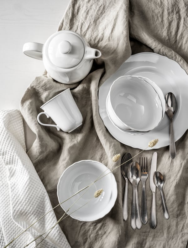 Vitt porslin med tallrikar, tekanna, kopp och bestick placerade på en beige linneservett.