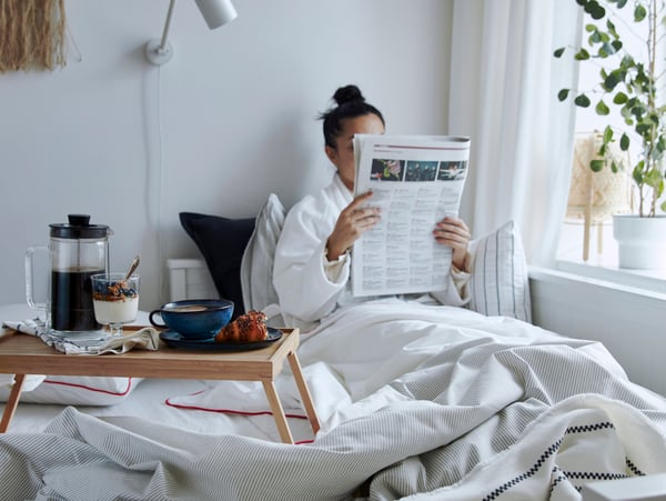 En person sitter i en säng med en sängbricka med frukost och läser en tidning.