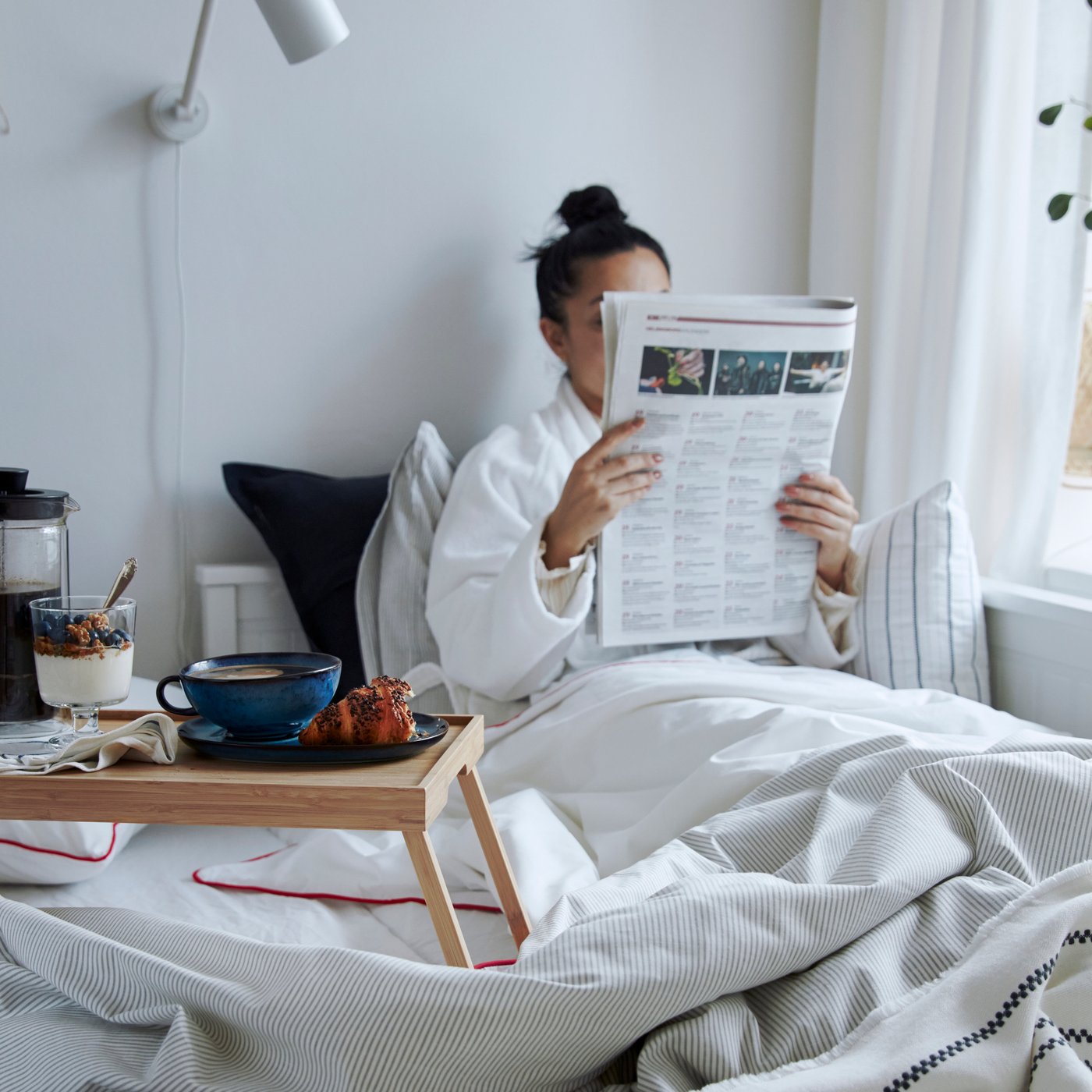 Een persoon in een witte badjas geniet van ontbijt op bed, leest een krant, met een IKEA KLIPSK bedtafel waarop een IKEA UPPHETTA cafetière, een blauwe mok en een yoghurtparfait staan, alles op een bed met een wit/donkergrijze IKEA STINAMAJ plaid.