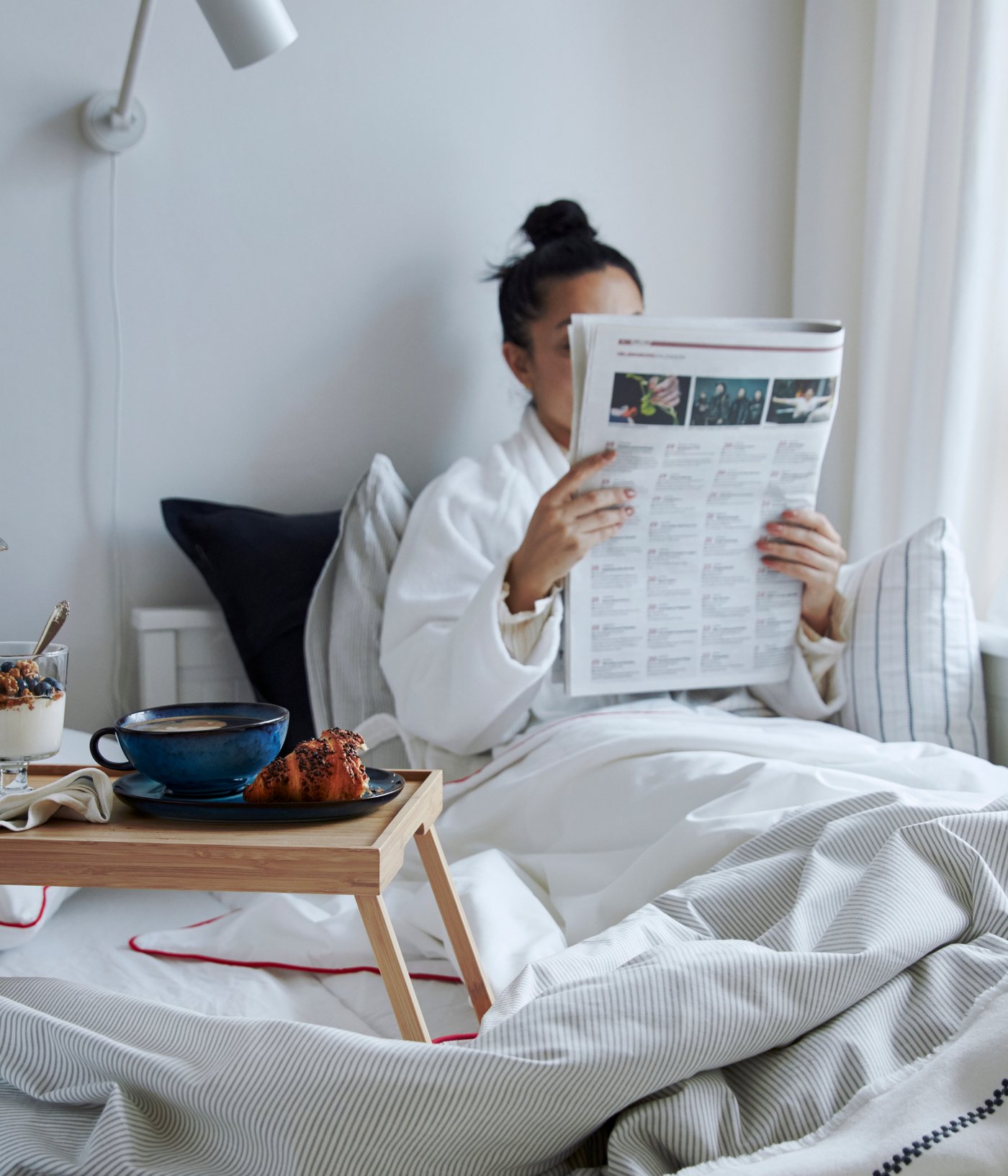 Een persoon in een witte badjas geniet van ontbijt op bed, leest een krant, met een IKEA KLIPSK bedtafel waarop een IKEA UPPHETTA cafetière, een blauwe mok en een yoghurtparfait staan, alles op een bed met een wit/donkergrijze IKEA STINAMAJ plaid.