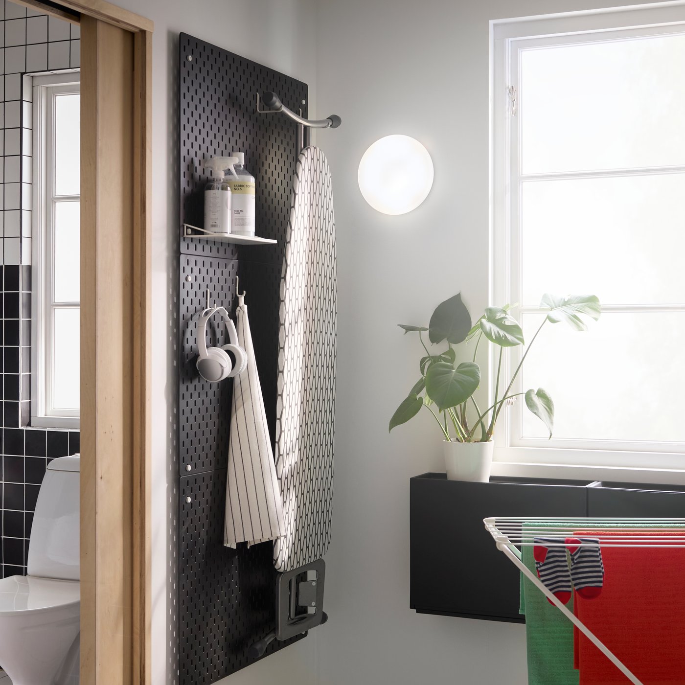 A black SKÅDIS pegboard on a bathroom wall displays an iron board, headphones and laundry soap. Sunlight from a window.
