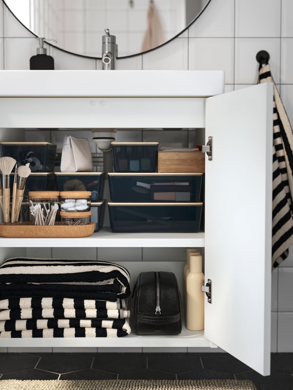 A bathroom vanity with open doors has glass jars with cork lids and white and black striped towels stored on the shelves.