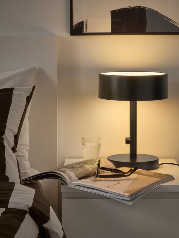 A modern, curved anthracite-coloured NYMÅNE table lamp stands on a bedside table, illuminating an open magazine.