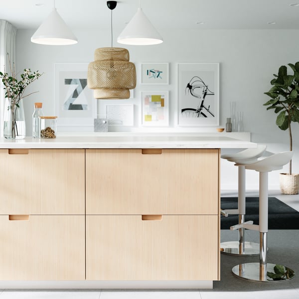 Modernt kök med ljusa träskåp, vit bänkskiva, tre vita barstolar, hänglampor och växt i hörnet.