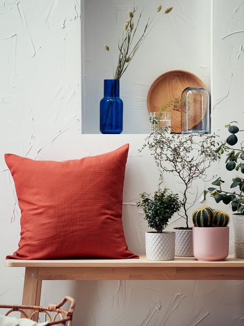 Un salon dans lequel on retrouve une housse de coussin orange, une table, des cache-pots, des plantes d’intérieur vertes, un vase bleu, un vase en verre clair et des objets décoratifs.