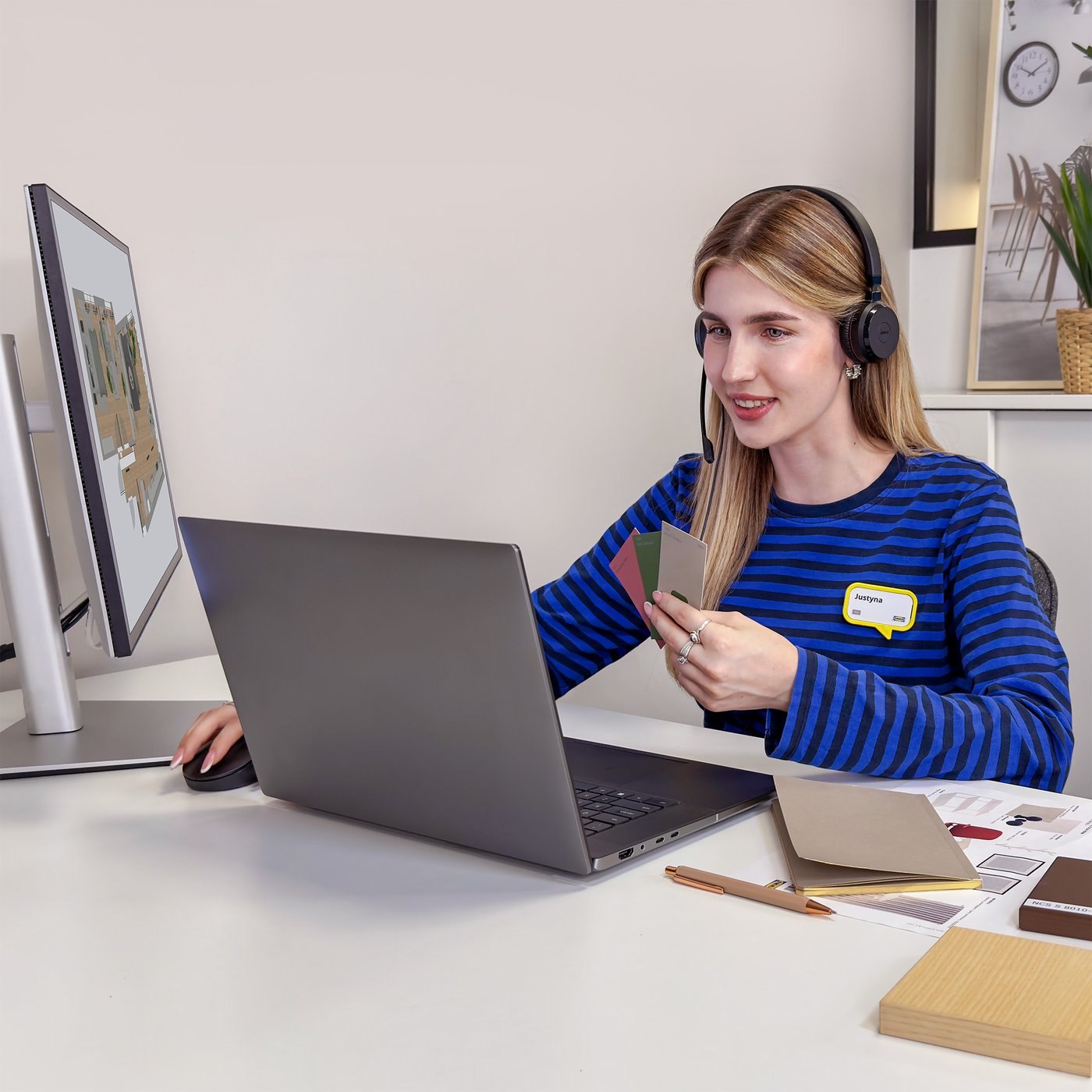 Projektantka IKEA siedząca przed laptopem przy biurku w niebieskiej koszulce z zestawem słuchawkowym na głowie. Prowadzi zdalnie usługę projektowania wnętrza i trzyma próbki kolorów w jednej ręce.