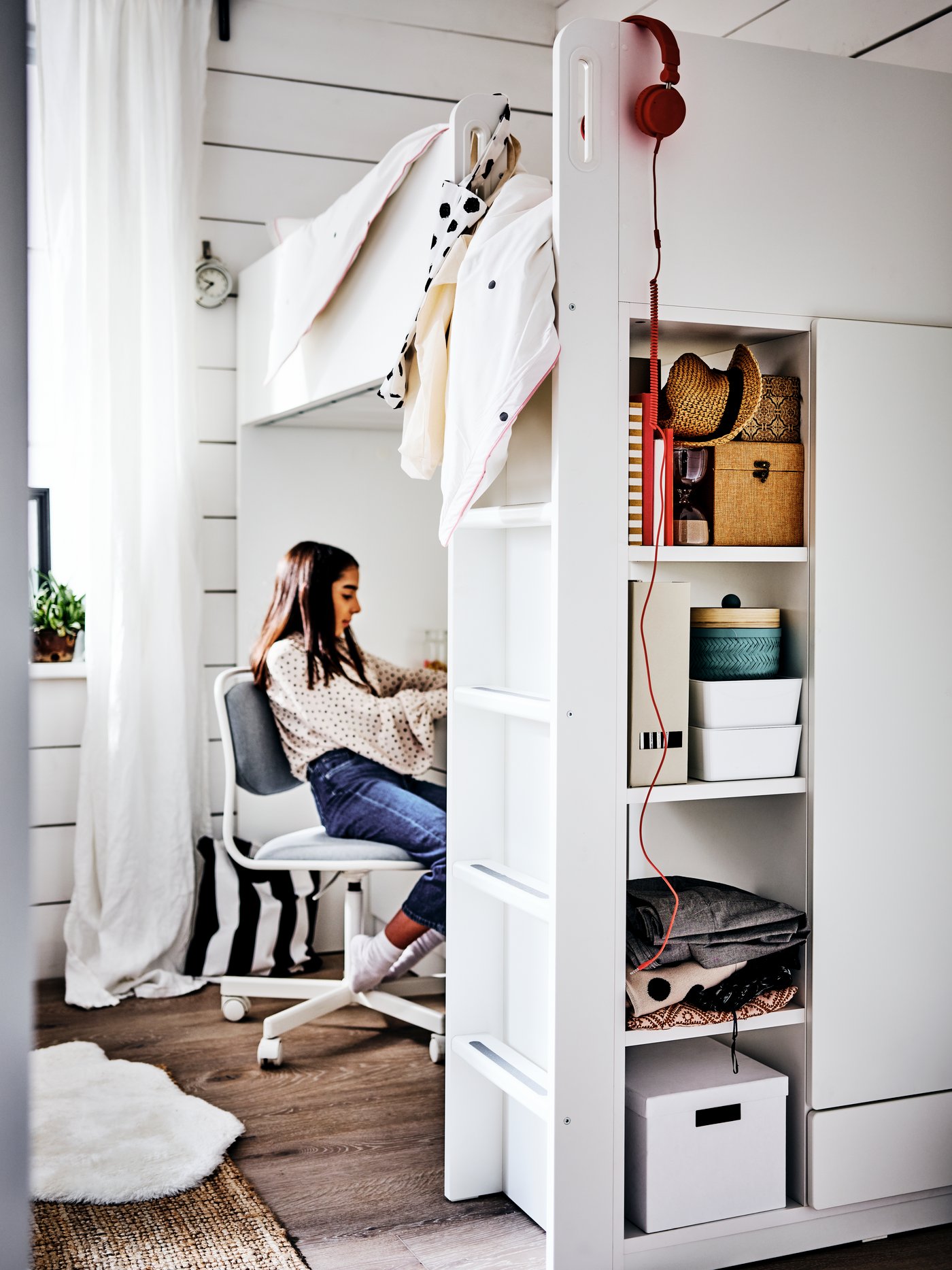 A young woman sitting on an ÖRFJÄLL chair at a desk under a SMÅSTAD loft bed. Boxes are stored on shelves in the bed frame.