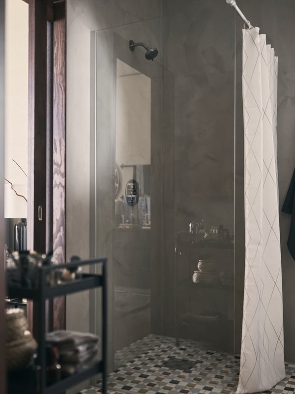A walk-in BROGRUND shower with a tiled floor that’s screened with a BASTSJÖN shower curtain, and next to a NISSAFORS trolley.