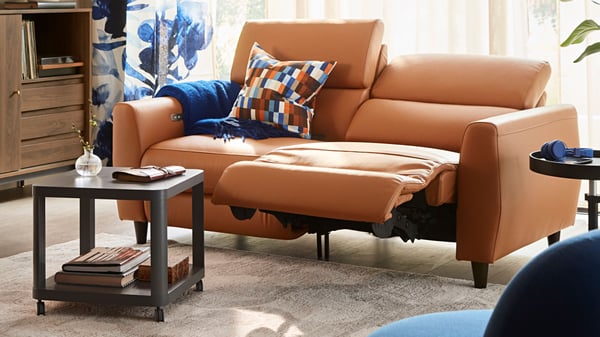 Soggiorno moderno con divano a due posti RULLERUM in pelle marrone con recliner elettrico reclino, affiancato da un tavolino e accessori eleganti.