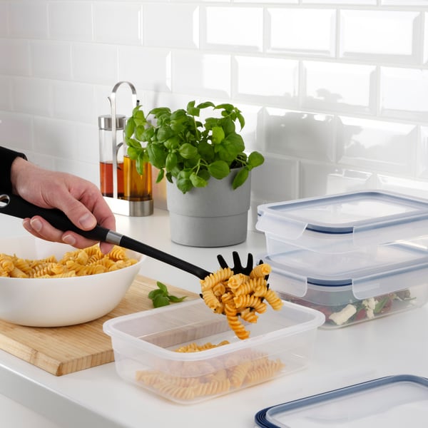 A stack of four clear, rectangular food storage containers with blue lids. From top to bottom, the containers are filled with green leafy vegetables, red cherries, green grapes, and yellow apricots. The containers are placed on a kitchen counter with a blurred background that suggests a home kitchen setting.
