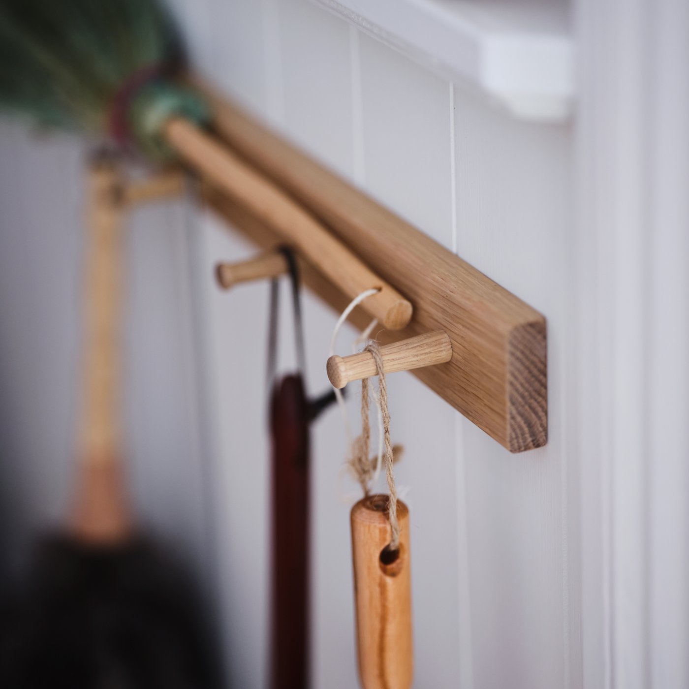 A section of a white wood-panel wall with a HÖVOLM oak six-knob rack holding various brushes and cleaning accessories.