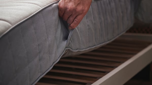 Une main soulève un matelas à ressorts ensachés VALEVÅG ferme/bleu clair posé sur un cadre de lit.