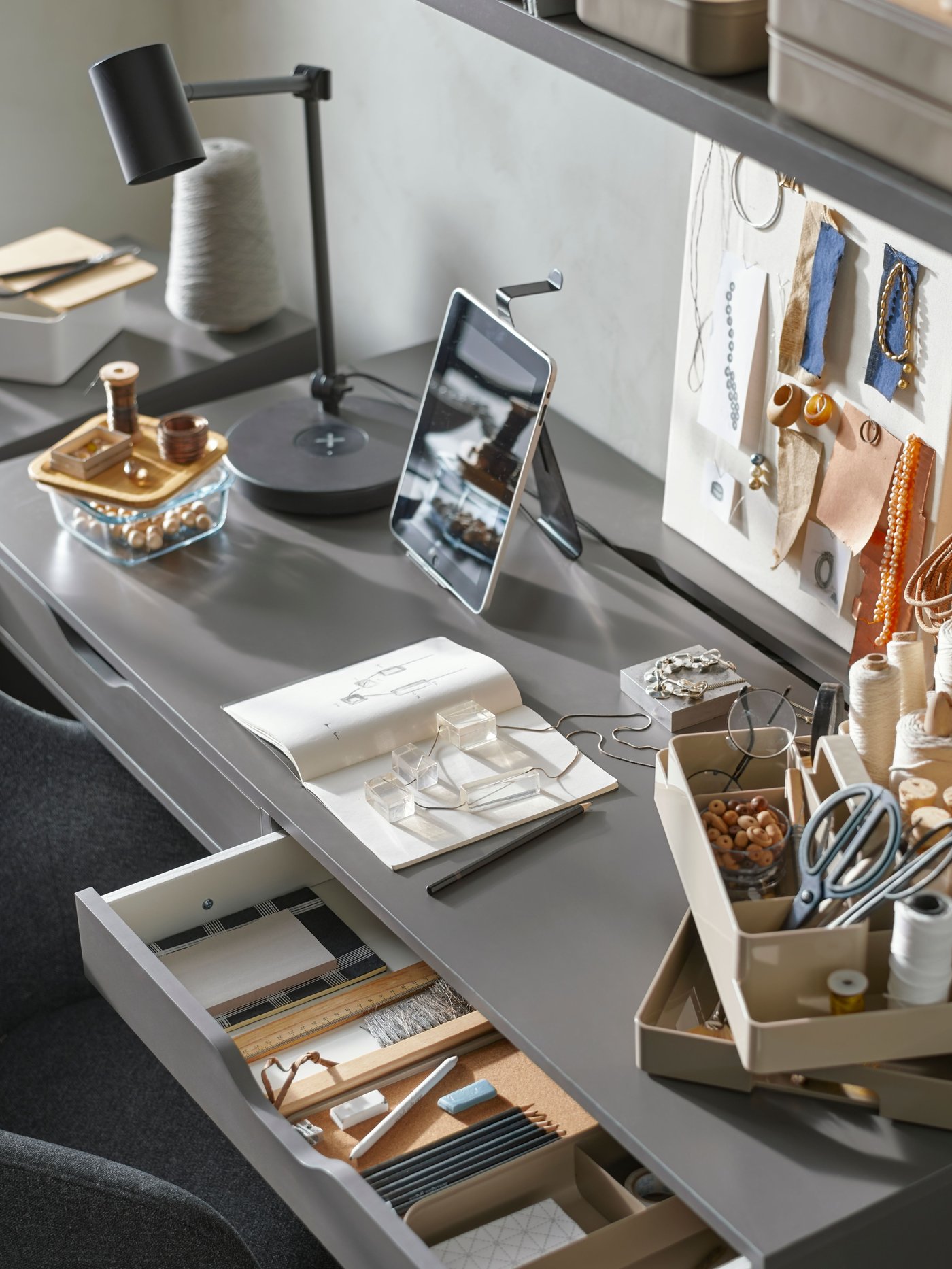 Un bureau ALEX gris foncé avec deux tiroirs, dont un à moitié ouvert. Un tableau d'inspiration, un carnet de croquis, du matériel de loisirs créatifs et un organiseur de bureau.