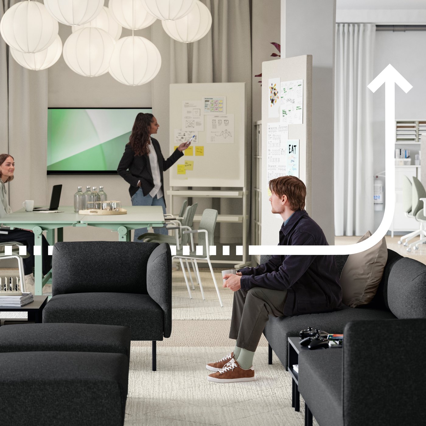 Ein junger Mann sitzt in einem Loungebereich, der aus mehreren dunkelgrauen LILLEHEM Sofamodulen und TUNSTA Tischen besteht, dahinter befindet sich ein Besprechungsbereich mit Whiteboard.