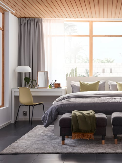 A white MALM bed and dressing table with a MORABO footstool stand in front of a large window, on a large grey STOENSE rug.