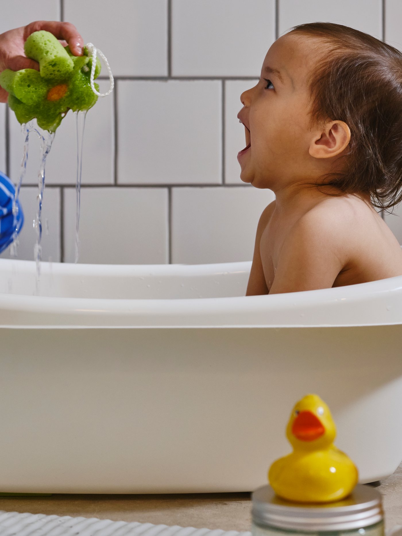 Ein Kleinkind sitzt in einer LÄTTSAM Babybadewanne und lacht, weil eine erwachsene Person Wasser aus einem grünen Schwamm spritzt.