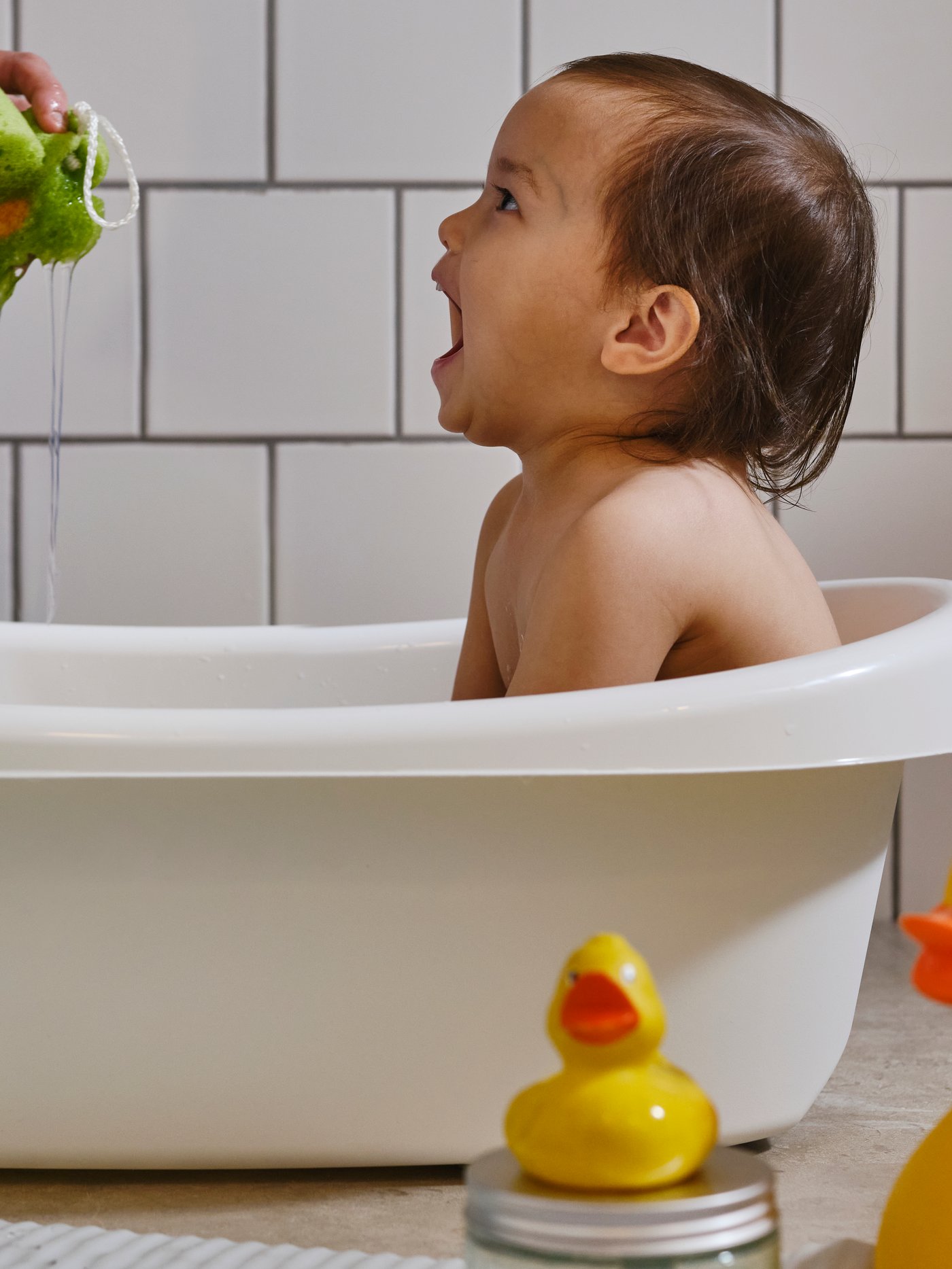 Et lille barn sidder i et hvidt LÄTTSAM babybadekar og griner, mens en voksens hånd klemmer vand ud af en grøn svamp.
