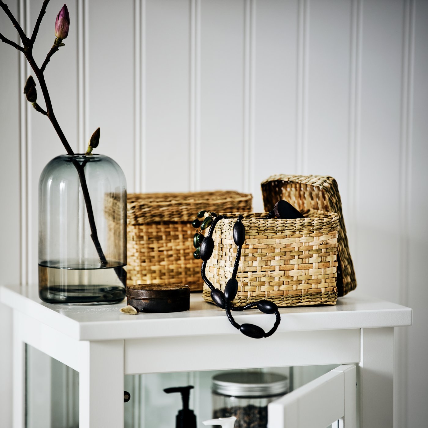 Two seagrass LURPASSA boxes by a vase on top of a HEMNES high cabinet. One box is open with a necklace hanging out.