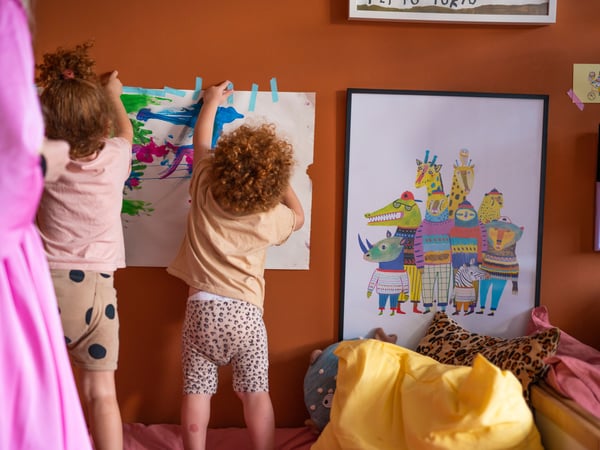Children taping a drawing on the bedroom wall next to other artwork.