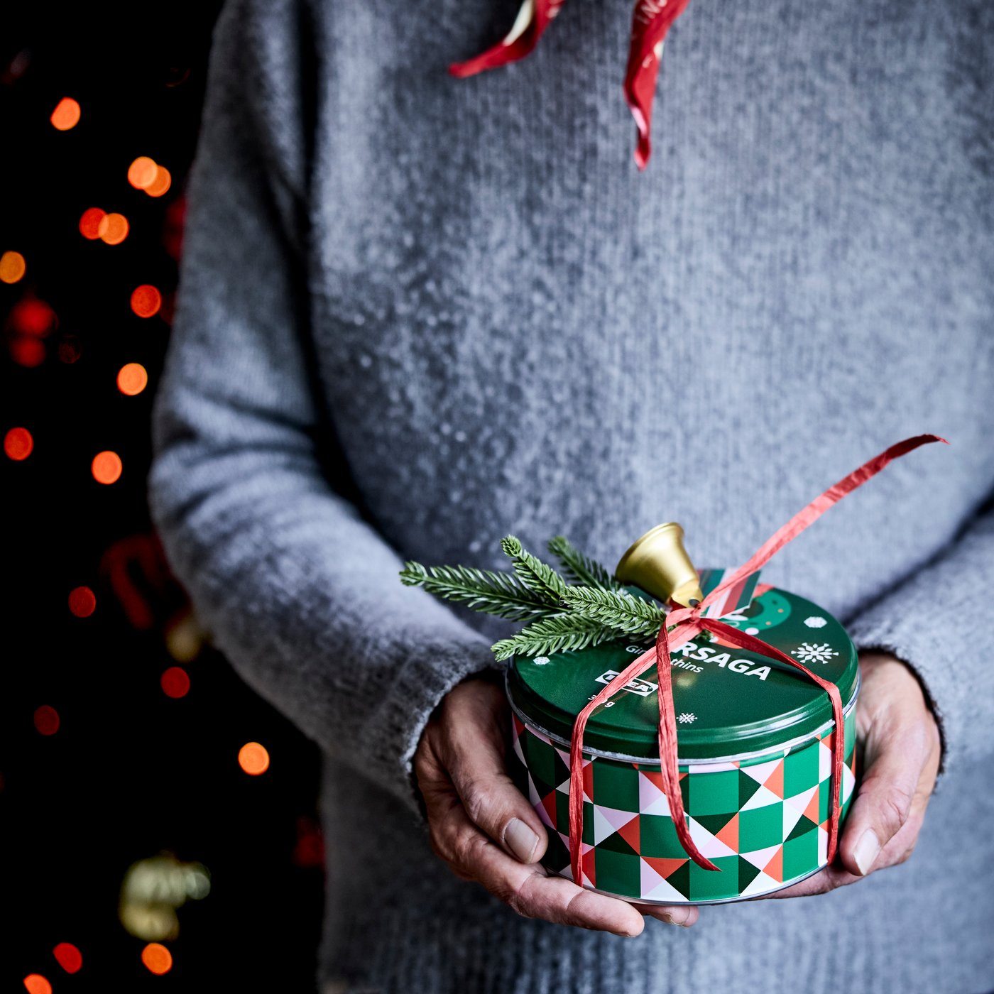 Person håller en grön rund presentburk med rött band och dekorativa grankvistar, framför en bakgrund med julbelysning.