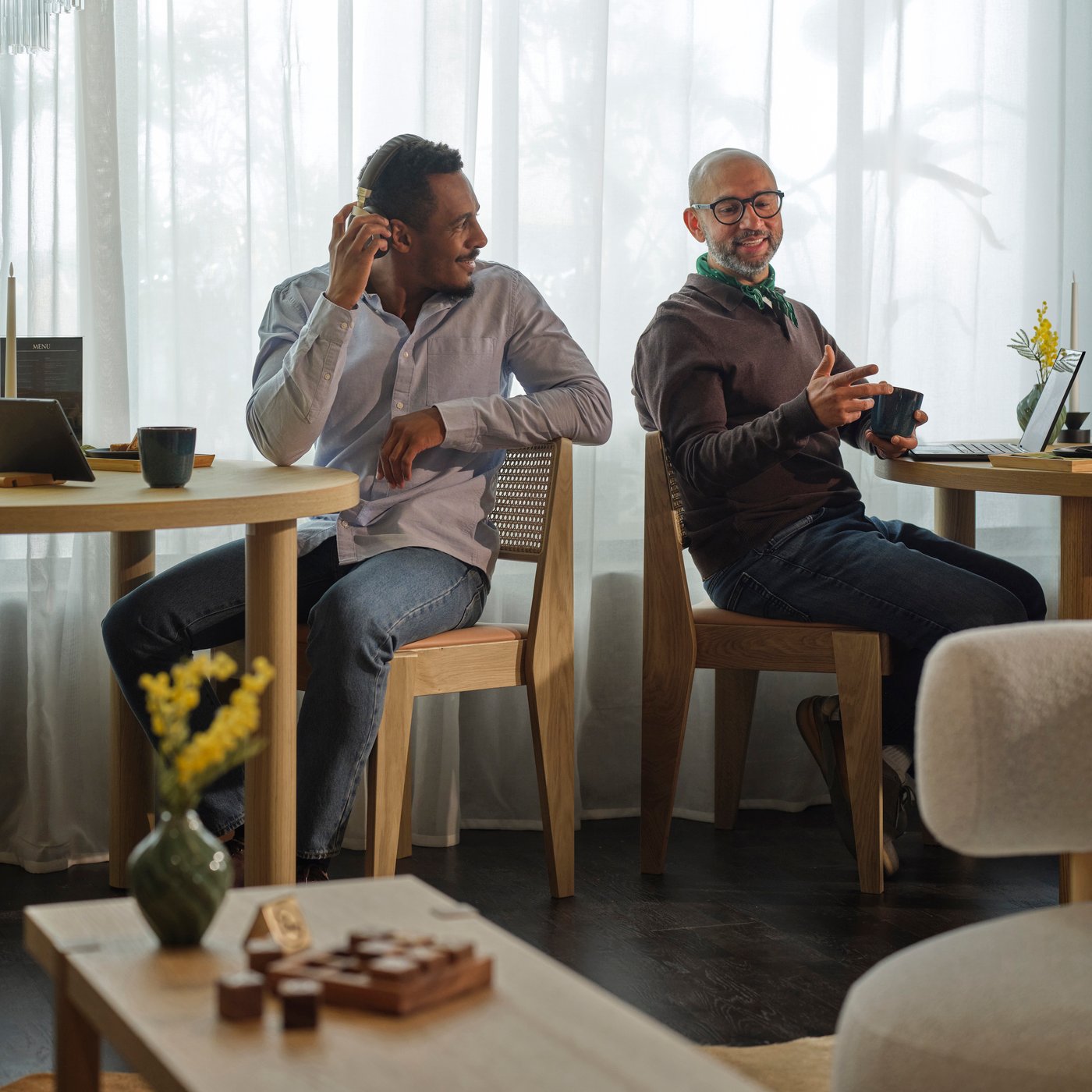 Två personer sitter vid separata bord, håller muggar och pratar i en ljus lounge med skira gardiner och trämöbler.