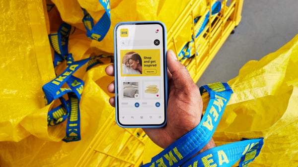 Smartphone with the IKEA app open in front of yellow IKEA shopping bags with blue handles – displaying user profile, text, and navigation icons