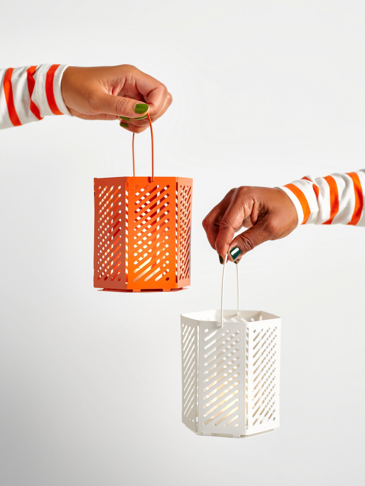 Two hands appearing from different sides, holding two VÅRSYREN lanterns for tealight, a white one and a brown-orange one.