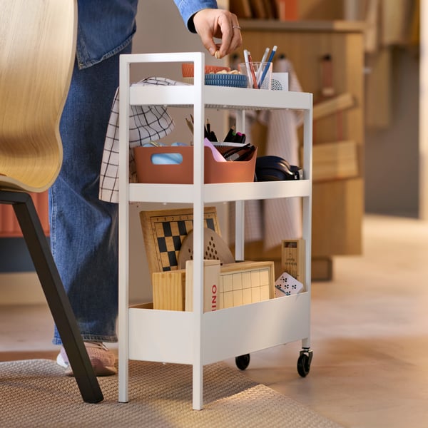 A white NISSAFORS trolley has different items stored on it and a person next to it is taking a nut from a bowl on a shelf.