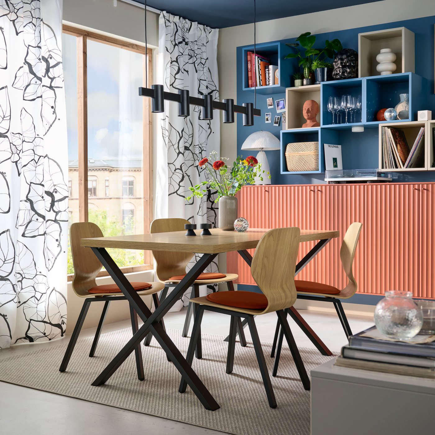 A room with large windows has a SKÅLSTA dining table with X-shaped legs and four chairs in oak veneer with black-grey legs.