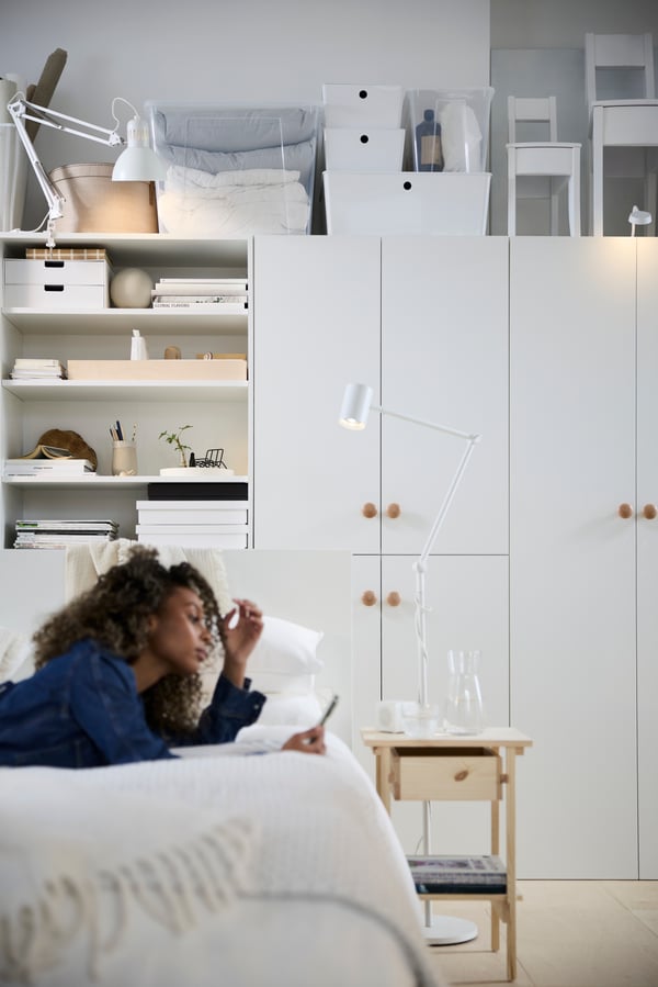 En person kopplar av på en säng i ett multifunktionellt bostadsutrymme med vita skåp och hyllor.