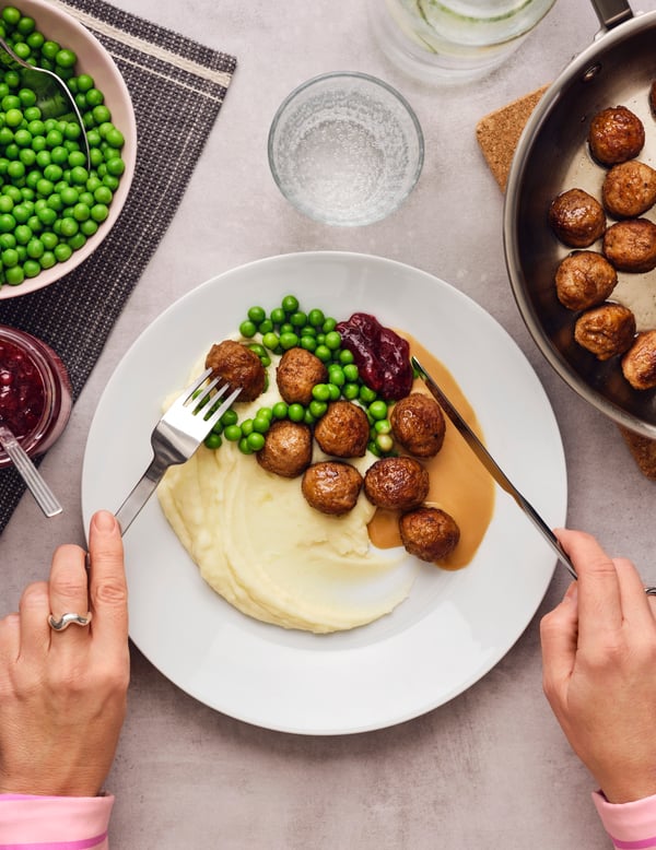 A white plate has a portion of meatballs, mashed potatoes and green peas, with other food dishes and a glass next to it.