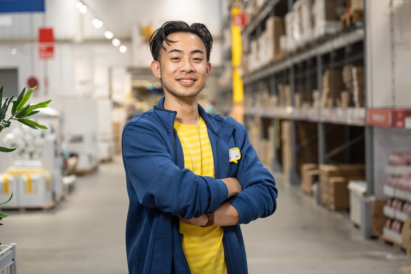 En person står med armarna i kors i en lagergång med hyllor och produkter i bakgrunden.
