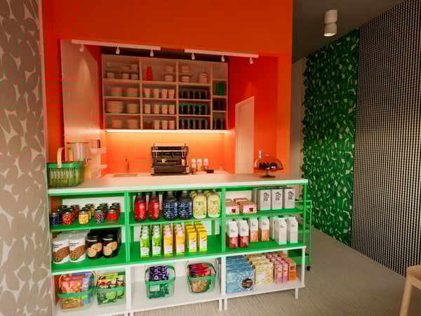 Teil des Cafés eines Hotels. Blick auf die Theke. Auf der Theke eine Kuchenplatte mit Glashaube Unter der Theke eine offene Aufbewahrung gefüllt mit verschiedenen Lebensmitteln. In der Rückwand eine Theke mit Kaffeeautomat und darübereine offene Aufbewahrung mit Geschirr.