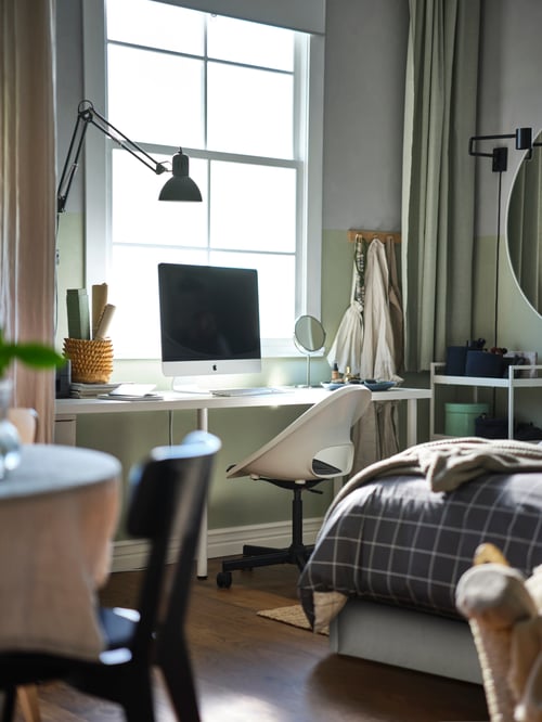 Ein Computer auf einem weißen LAGKAPTEN/ADILS Schreibtisch mit einem LOBERGET/MALSKÄR Drehstuhl mit Polster neben einem Bett unter einem Fenster.