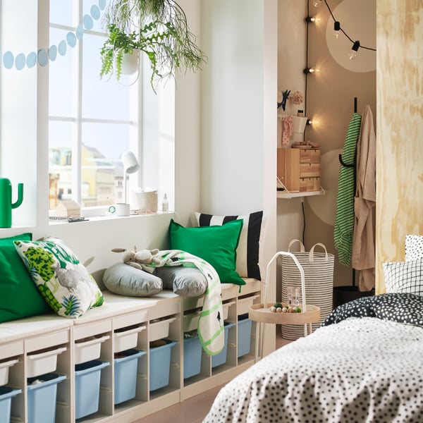 A cosy room featuring a TROFAST frame in white holding colourful storage boxes and with cushions on top under a sunny window.