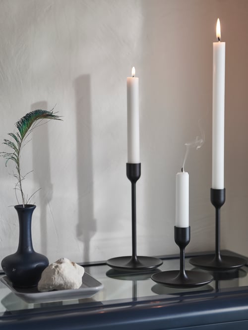 Prim-plan cu trei suporturi negre pentru sfeșnice care țin lumânări albe fără parfum lângă o vază albastră adâncă pe o tavă gri de copt.