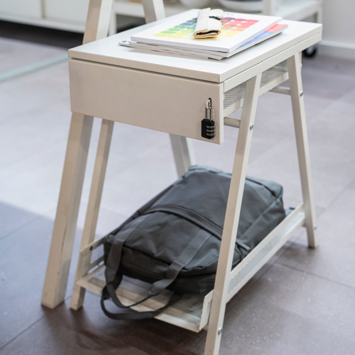 A white desk sits holding a gray backpack in an underneath compartment. 