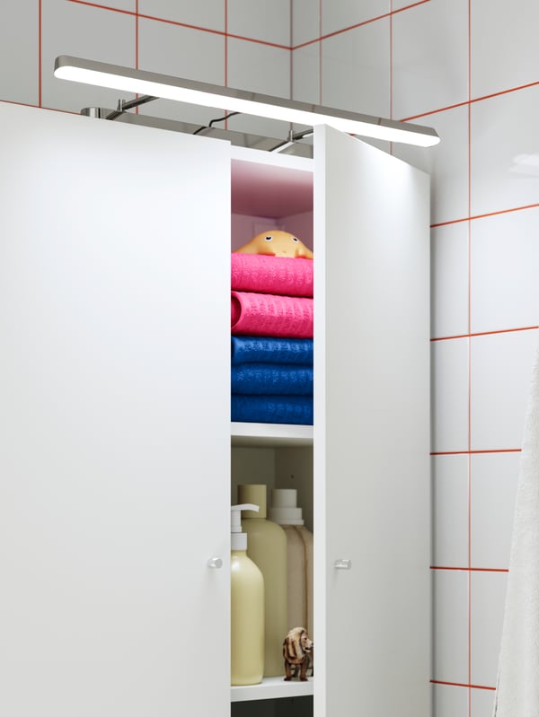 Open cabinet with FLASKPOST LED cabinet/wall lighting, displaying neatly stacked towels and toiletries in a tiled bathroom.
