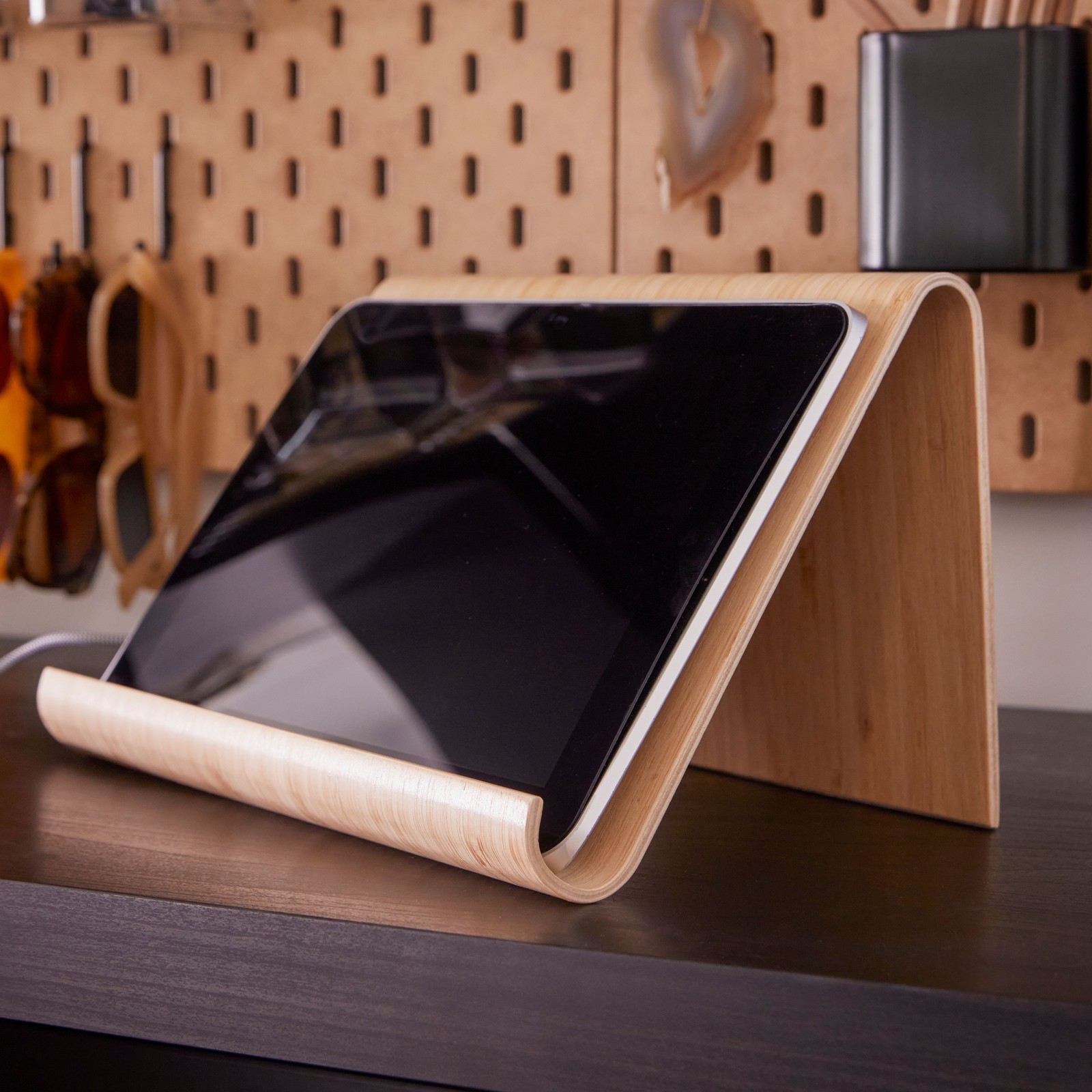 A horizontally placed tablet in a VIVALLA bamboo veneer tablet stand on a desk by a wall with a pegboard attached to it.