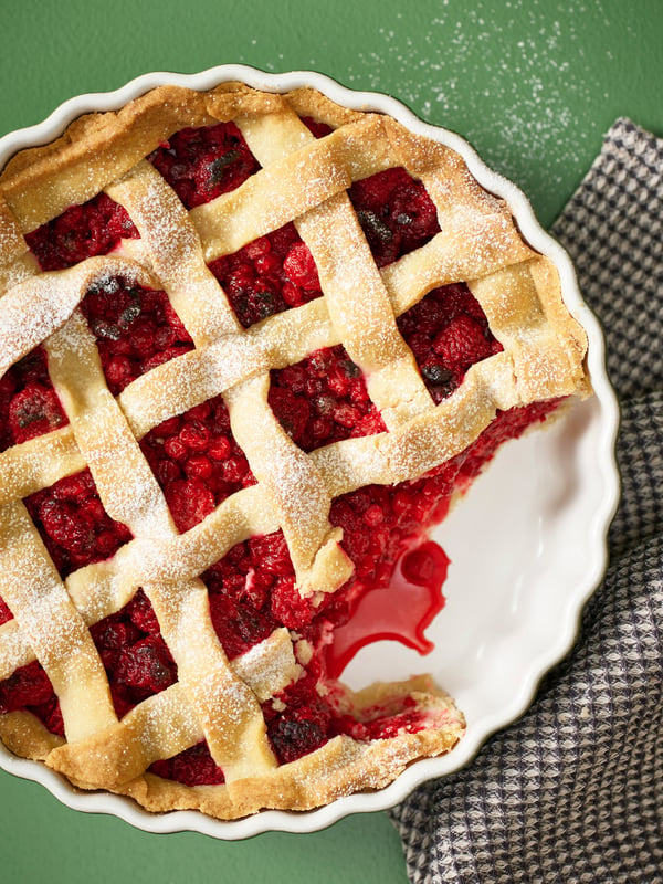 A berry pie in a white/dark green NÄBBFISK pie dish is placed on a kitchen towel, with a piece taken out of the pie.